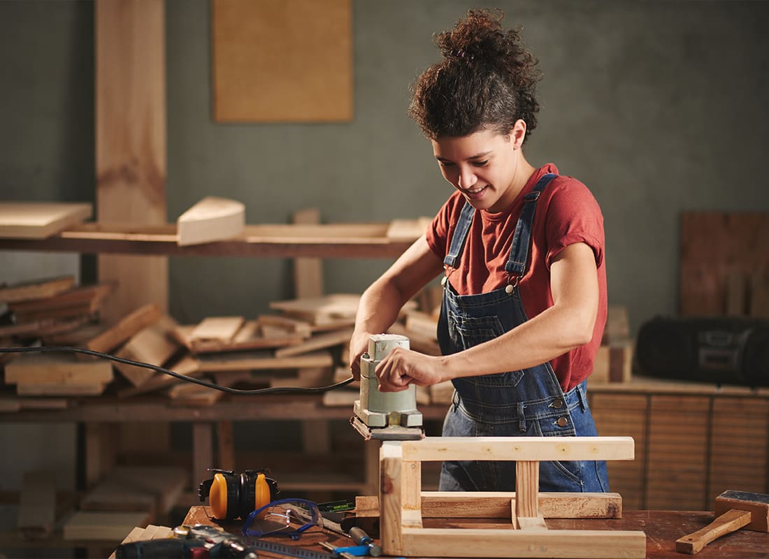 Insurance by Industry - Female Carpenter Sanding Down Wood in Her Workshop